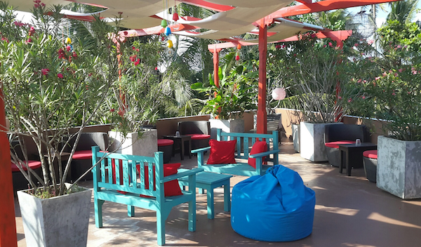 A close-up of a rooftop terrace with colorful tables, chairs and plants. Treetops can be seen in the background.