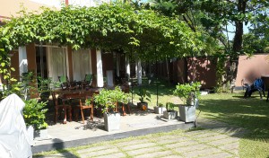 A picture of a pergola with table and chairs underneath. In the background guest house and garden.