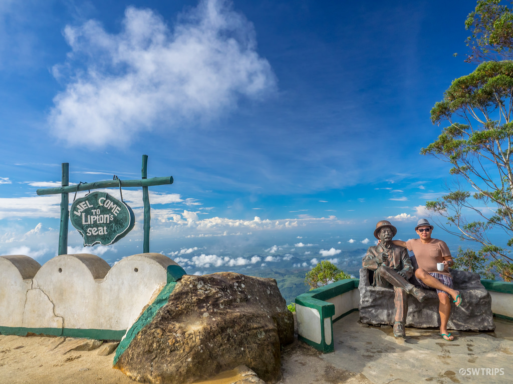 A picture of a bench with a man in bronze and a man in a hat, with a view of the landscape behind and a blue sky with clouds. The bench is called Lipton Seat and can be found in Haputale.