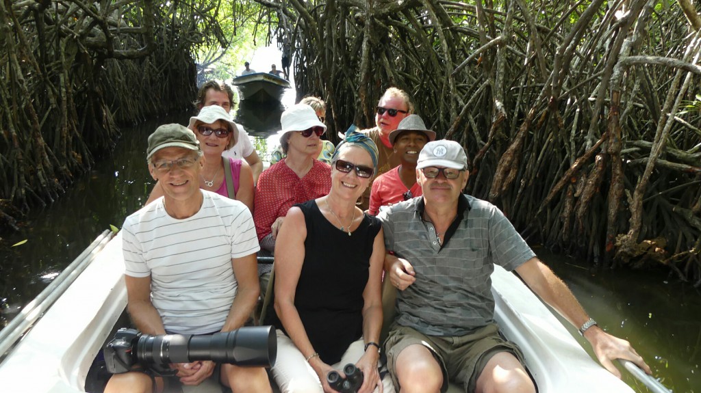 A close-up picture of nine elderly people in a small boat in a channel between mangrove roots. In the background a boat and sunlight in the opening to the canal.