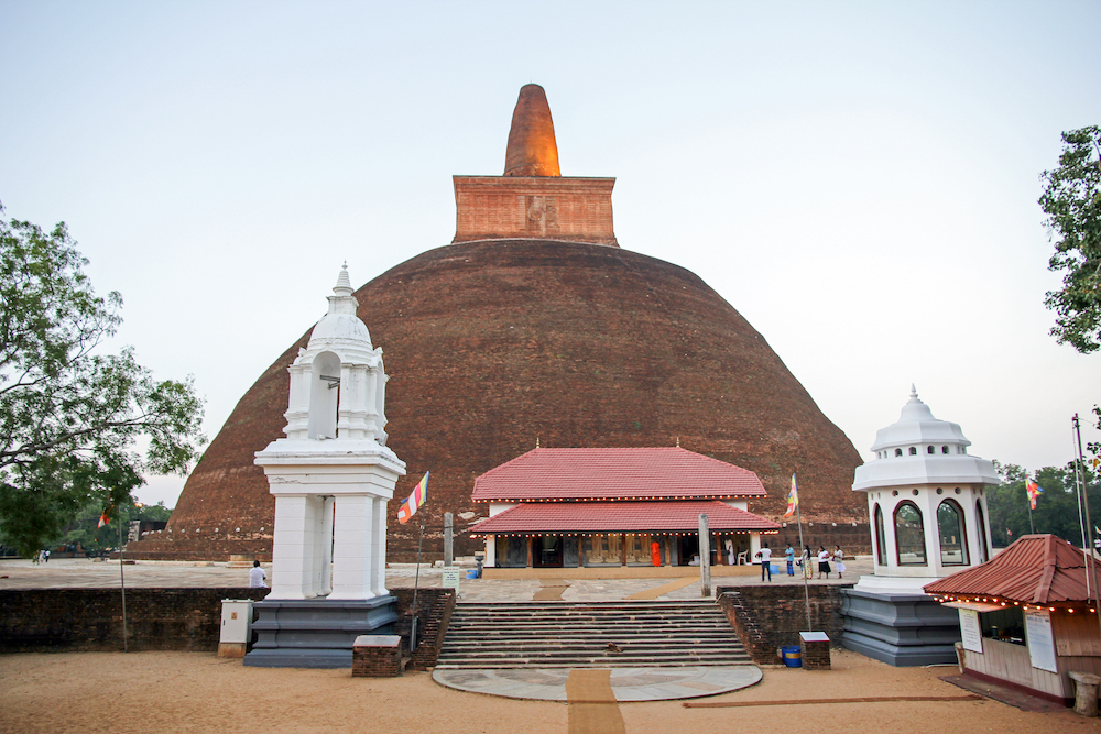 A picture of a large domed stone temple in Anuradhapura. A staircase in front and trees with sky in the background.