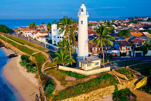 A distant shot picture of a white lighthouse standing on the ruins of a thick wall along the shore. In the background a city and blue sky.