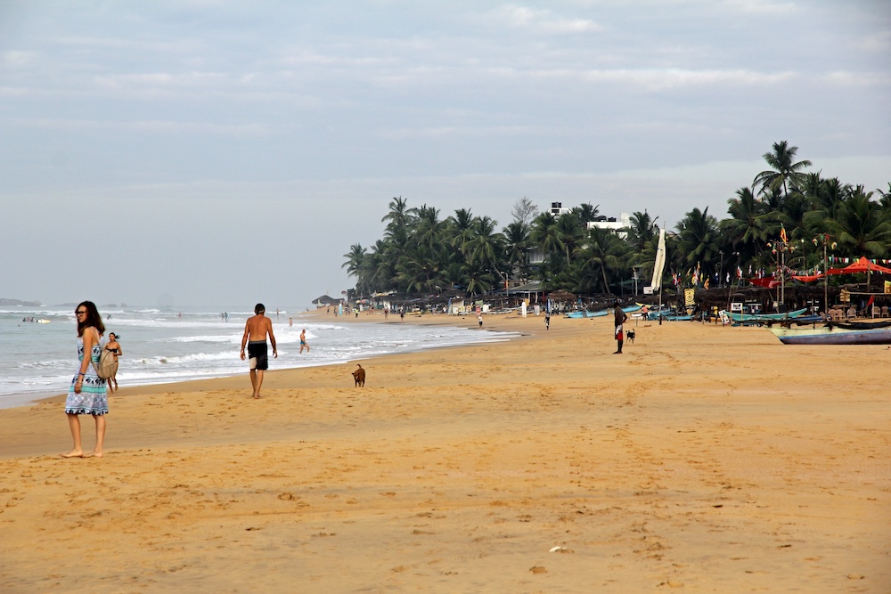 A picture of a wide sandy beach and a bit of the ocean, with small boats and palm trees in the background.