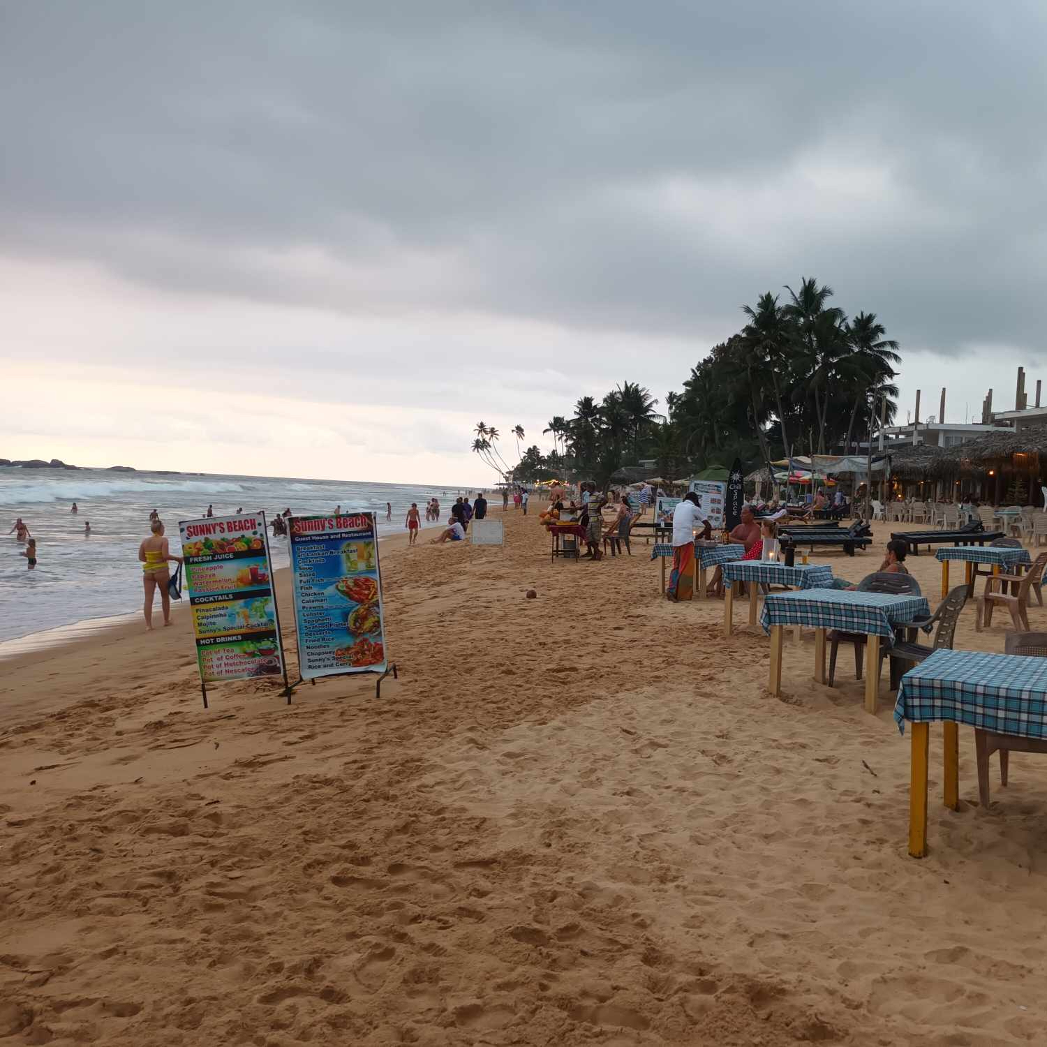 A picture of a beach and sea with tables and sunbeds. Trees and gray sky in the background.