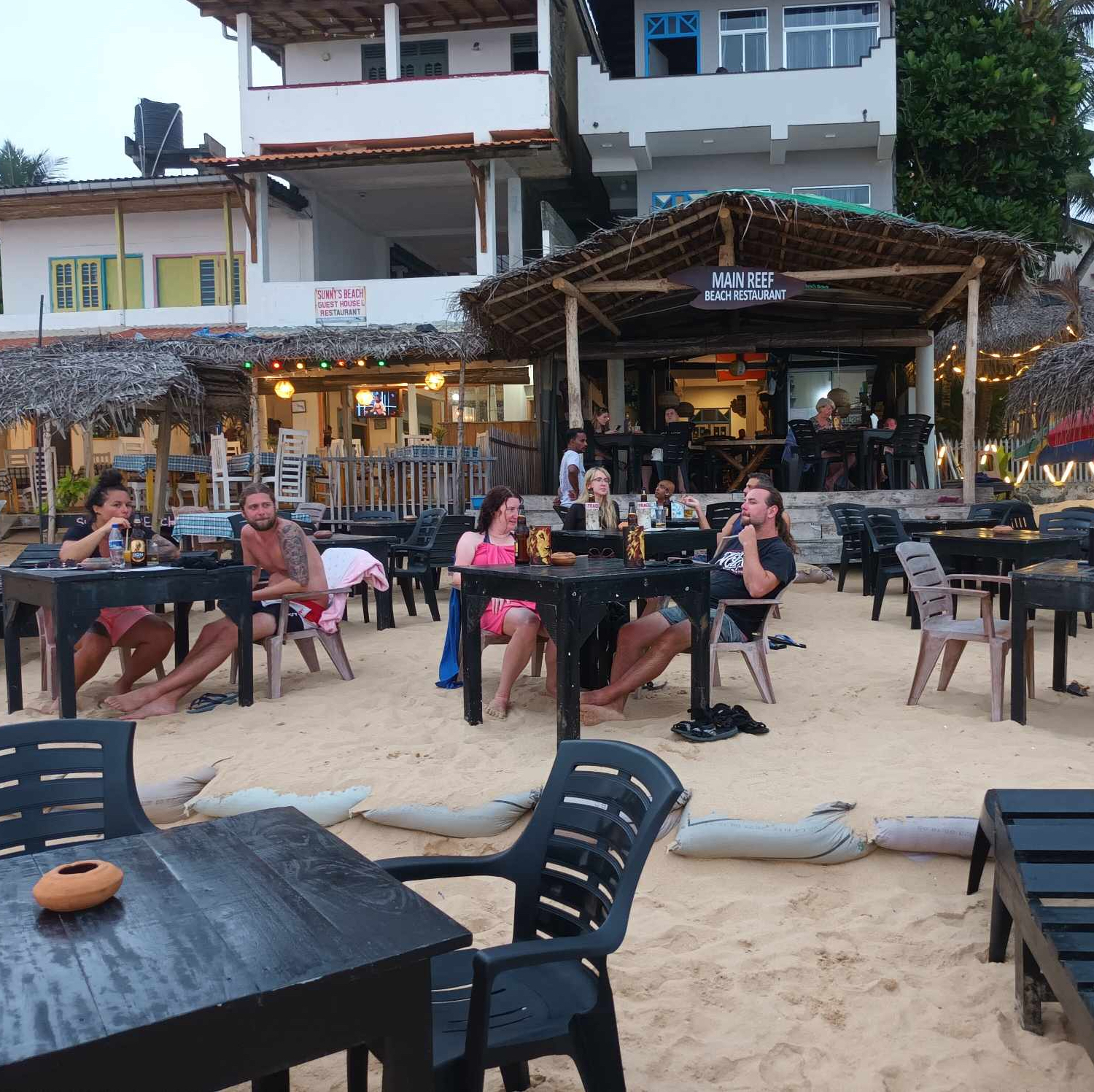 A picture of restaurants on the beach with tables and guests sitting in front.